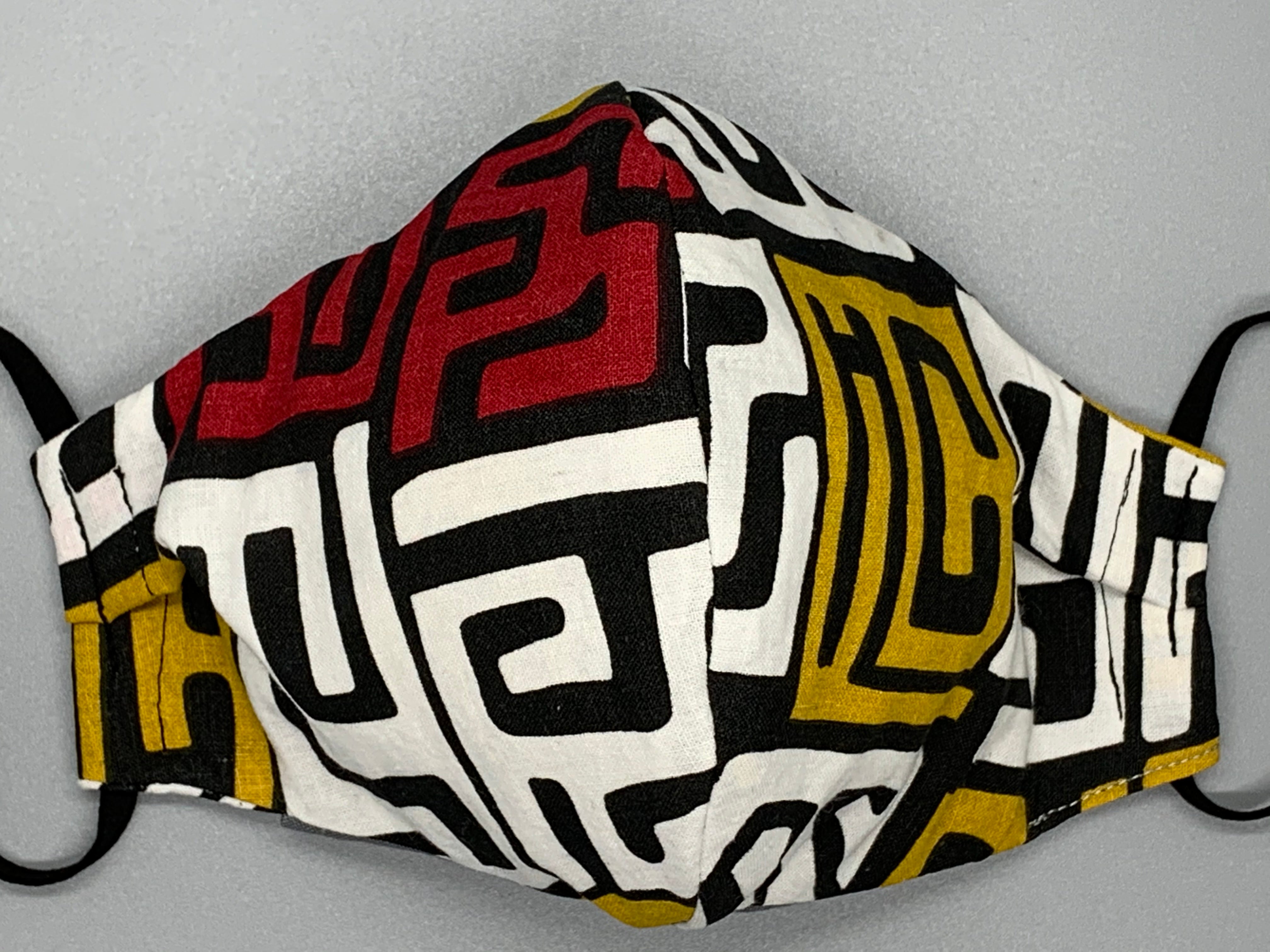 Handmade Triple Layer Cotton Facemask. White, Red, and Mustard symbols maze across black cotton. Gray filter pocket. Overhead/Back of Head cotton loop from same fabric. Adjustable Wood Bead for perfect fit. This mask has no nose wire-the adjustable overhead loop gives you a snug fit. Cut from one cloth-meaning fabric is same and where your pattern lies may vary. non-returnable/non-refundable. non medical grade. not a replacement for medical grade personal protective equipment (PPE), surgical masks, N95s, or