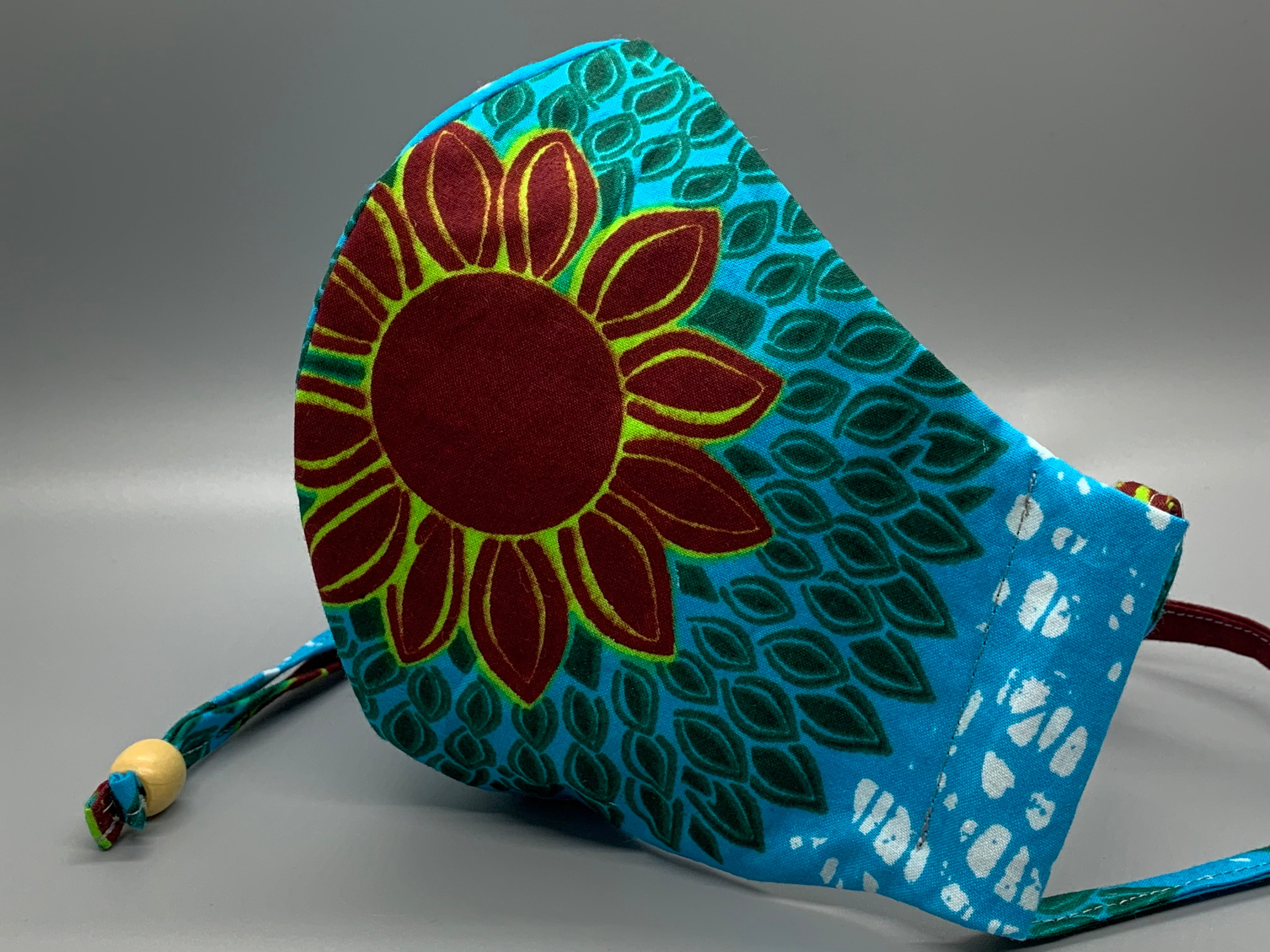 Lay down in a sunflower field on a cloudless day. This African wax cloth features maroon sunflower, green petals, bright blue, and white. Gray filter pocket. Overhead/Back of Head cotton loop from same fabric. Adjustable Wood Bead for perfect fit. This mask has no nose wire-the adjustable overhead loop gives you a snug fit.