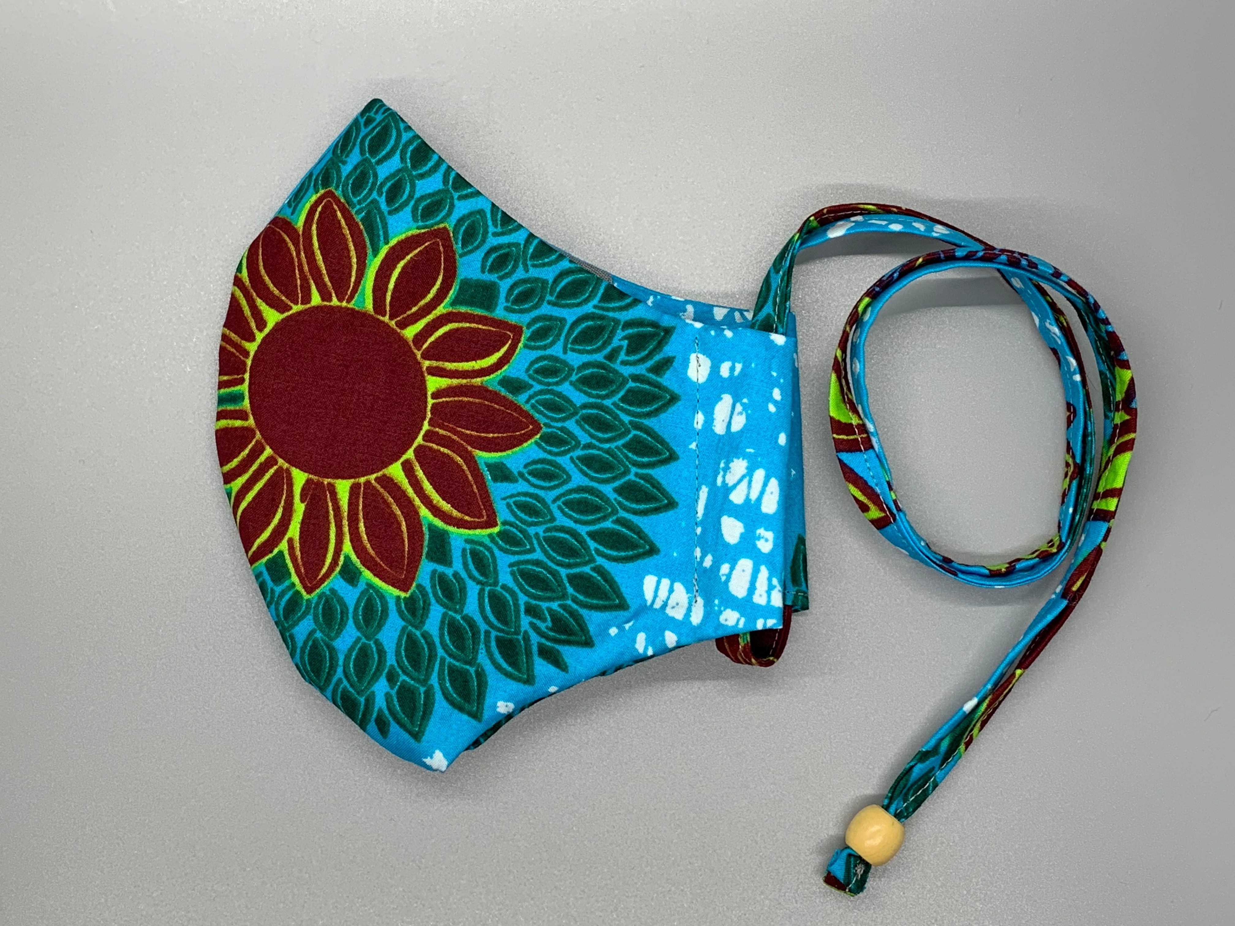 Lay down in a sunflower field on a cloudless day. This African wax cloth features maroon sunflower, green petals, bright blue, and white. Gray filter pocket. Overhead/Back of Head cotton loop from same fabric. Adjustable Wood Bead for perfect fit. This mask has no nose wire-the adjustable overhead loop gives you a snug fit.